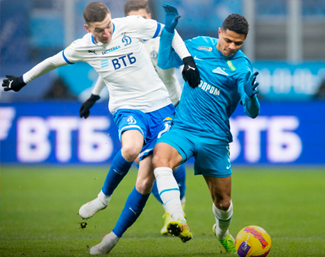 Два футболиста борются за мяч на поле. Игрок в бело-синей форме пытается отобрать мяч у соперника в синей форме. На заднем плане виден нечеткий фоновый баннер с логотипами