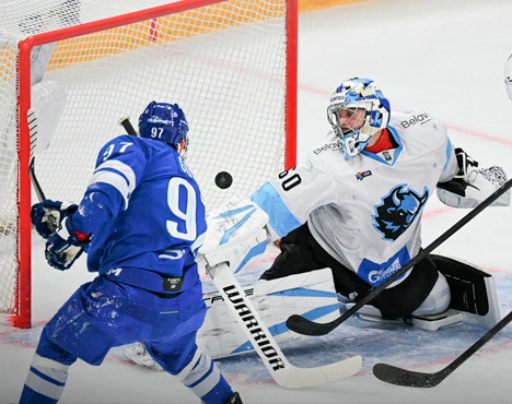 Напряжённый момент в хоккейном матче: игрок в синей форме атакует ворота, пытаясь забить шайбу, в то время как вратарь в белой форме делает попытку её отразить. Шайба находится в воздухе